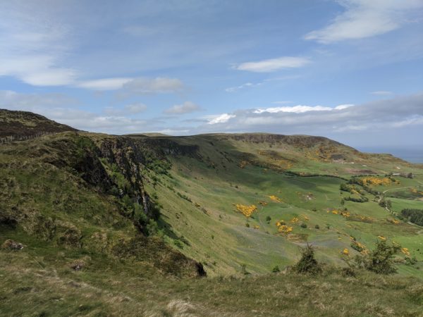 Looking North to Sallagh Braes, with the floor of the dramatic escarpment resplendent in the sunshine, lush green grass and small purple flowers